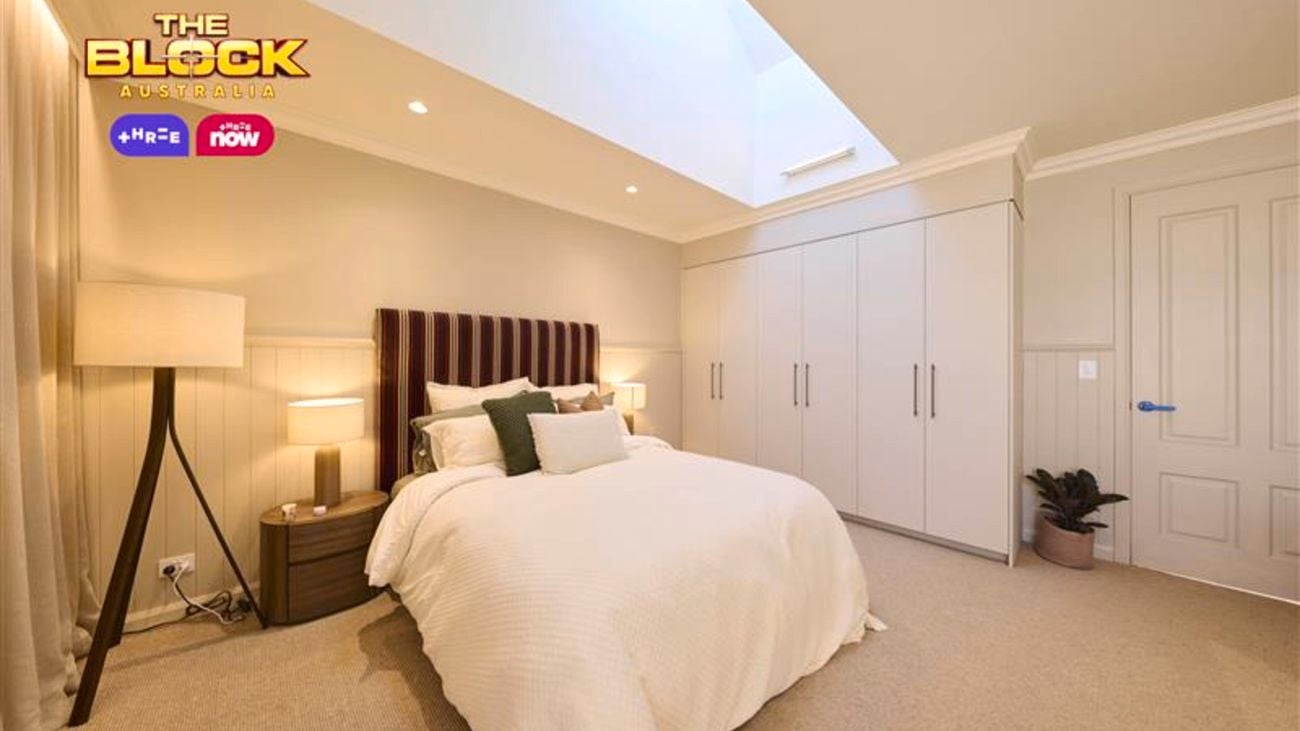 Cream bedroom with a double bed and a patterned bedhead, white linens and a lamp in the corner and a wardrobe opposite.