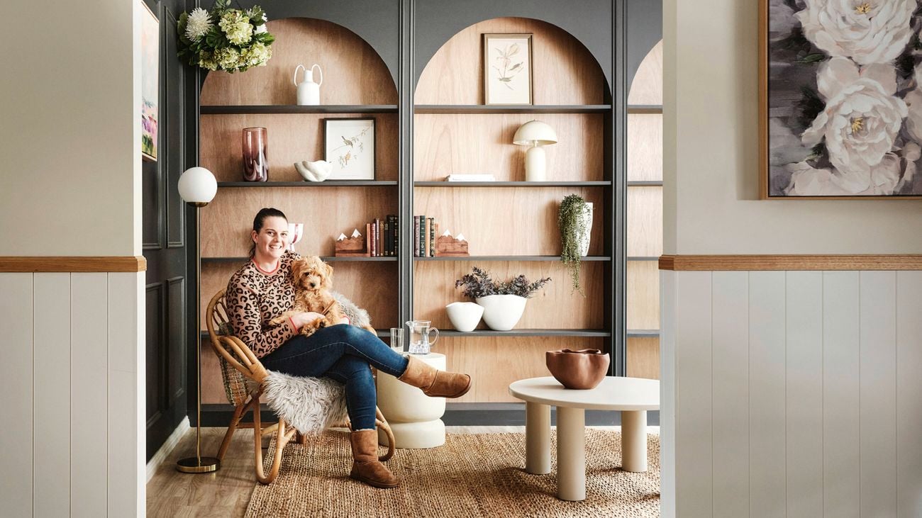 A woman sitting in a wicker chair with a small dog on her lap, in front of a large wooden bookshelf filled with decorative objects, alongside a coffee table and a textured rug.