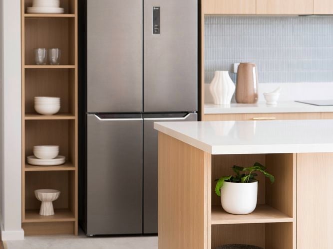 A light brown kitchen with a fridge, a white benchtop and shelving with crockery on it. 