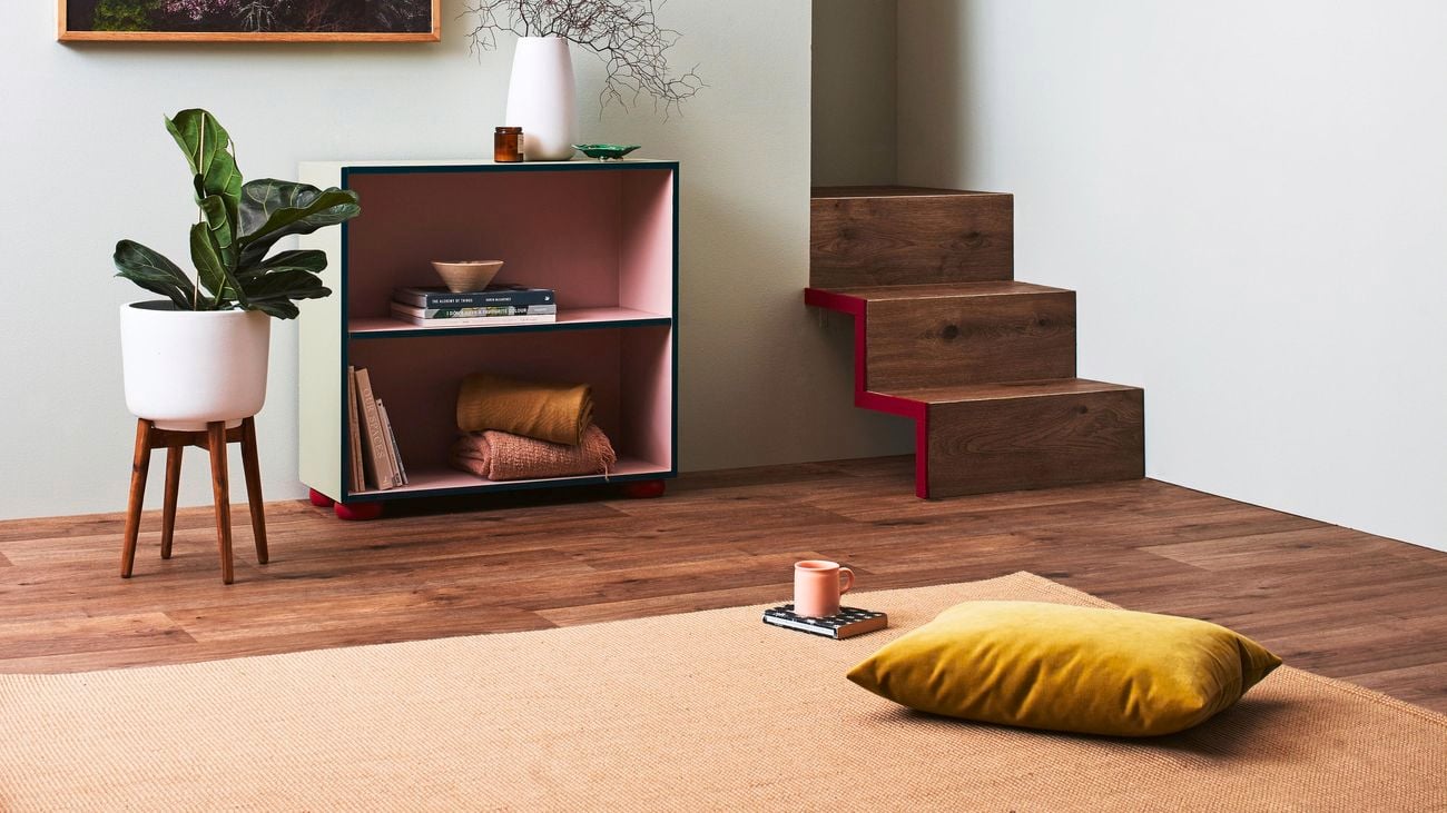 A lounge room with a cushion, pot plant, shelving unit and a painting on the wall.