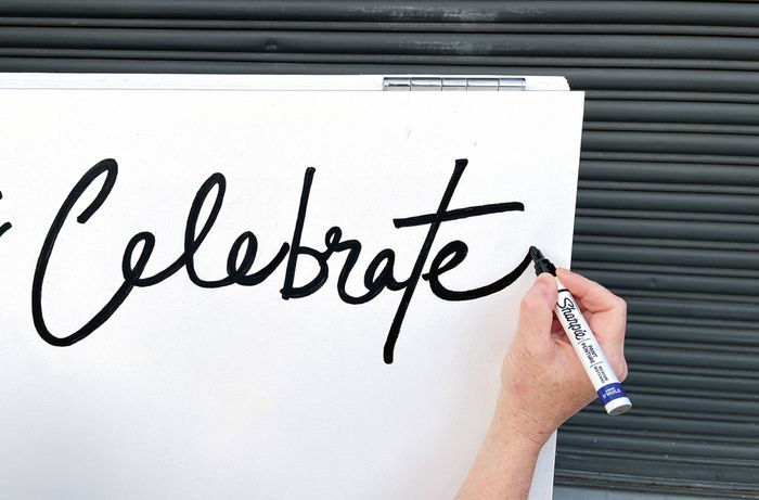 A hand using a black marker to write "Let's Celebrate" in script on a white-painted plywood panel. [[2609] MAK...me sign_V1 | Word]