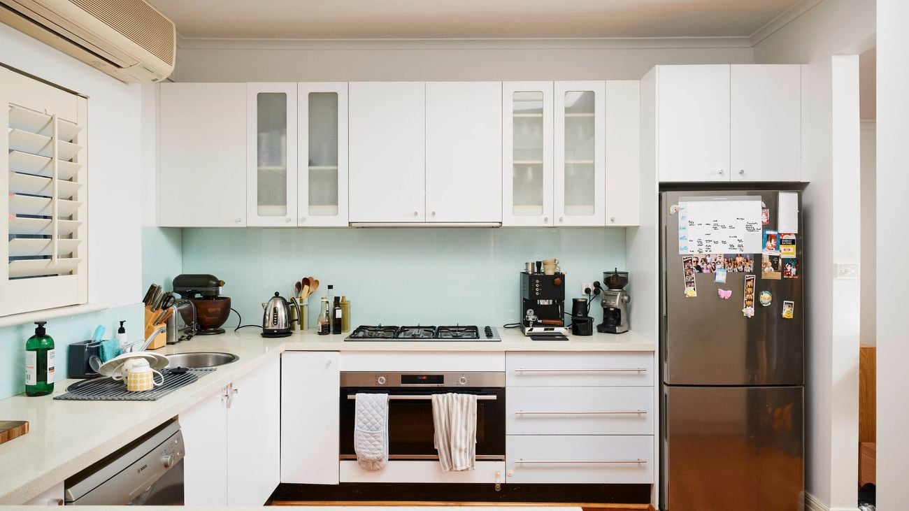 Modern white kitchen with overhead cabinetry and a glass splashback, featuring a built-in oven and gas cooktop, a stainless steel fridge with magnets, and various small appliances and utensils arranged along the benchtop.