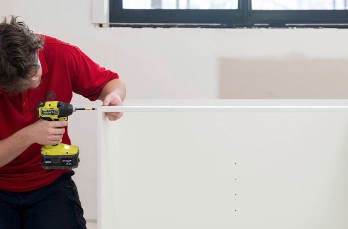 A Bunnings Team Member is using a power drill to add the top support rail across the cabinet frame. 
