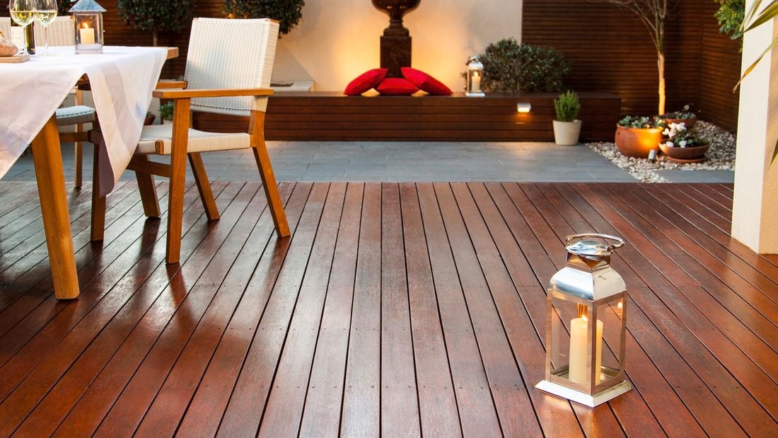 A stained deck with a nearby dining table and chair, and a lit lantern, overlooking a paved outdoor area with benches and cushions