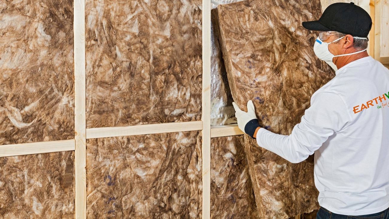 A person wearing gloves and a mask holding a wall batt of Earthwool insulation to install it in a wall.