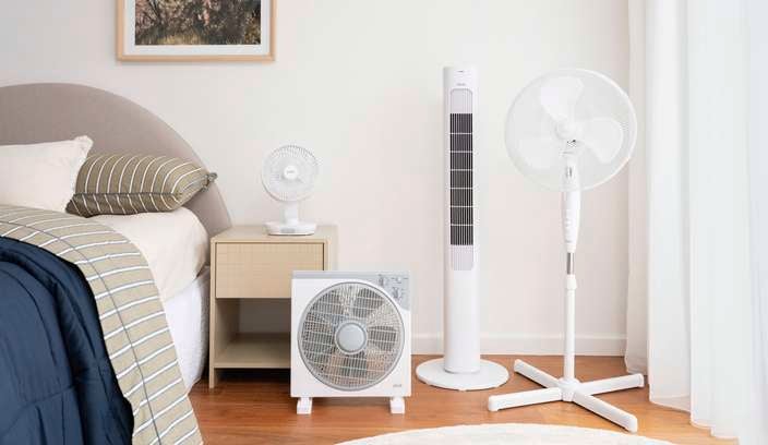 Collection of cooling fans including a desk fan, box fan, tower fan and pedestal fan arranged in a bedroom beside a bed and window.