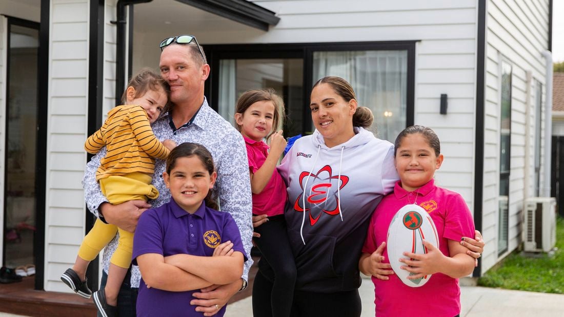 A group of adults and children stand together outside a modern white house. One adult holds a small child, while another adult stands beside several children. One child in a purple shirt stands with arms crossed, and another child in a bright pink shirt holds a rugby ball. The group appears to be posing for a family or community photo.
