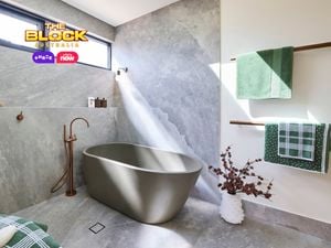 Modern bathroom with matte gray freestanding tub, stone-look gray tiles, wall-mounted faucet, green towels on wooden racks, and natural light from a high window. 