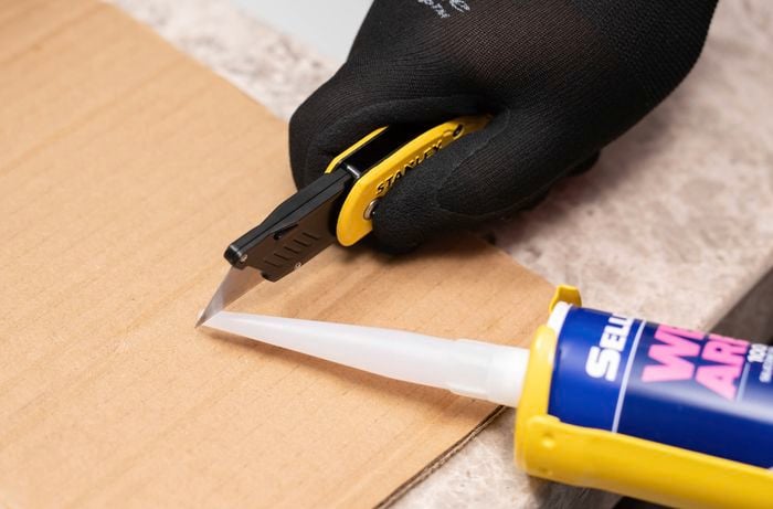A person is trimming the silicone nozzle at a 45-degree angle using utility knife for controlled sealant application. ​