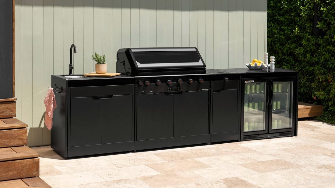 A black outdoor kitchen with a barbecue, sink and bar fridge in it, sitting on paving outside.