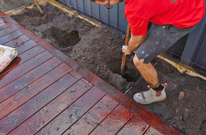 A person digging a hole between a deck and a paling fence