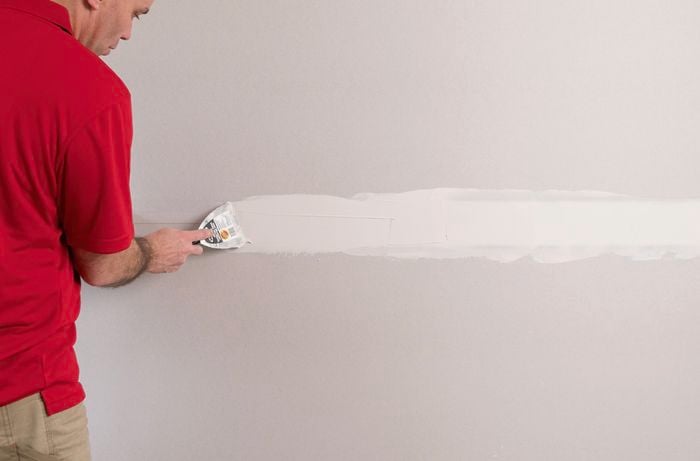 A person in a red T-shirt uses a plastering trowel to apply a thin layer of base coat compound over the tape.