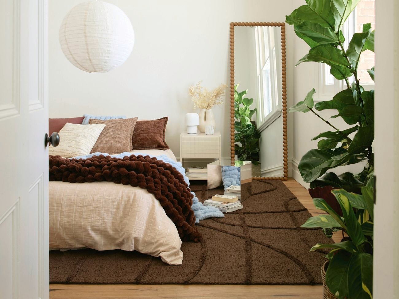 Styled bedroom featuring soft neutral bedding, chunky throw, textured brown rug, indoor plants, and decorative mirror creating a warm, contemporary space.