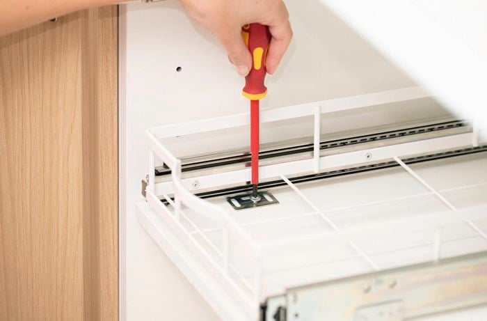 A person is using a screwdriver to tighten fixings on a pull-out drawer system after the rails have been positioned inside the cupboard