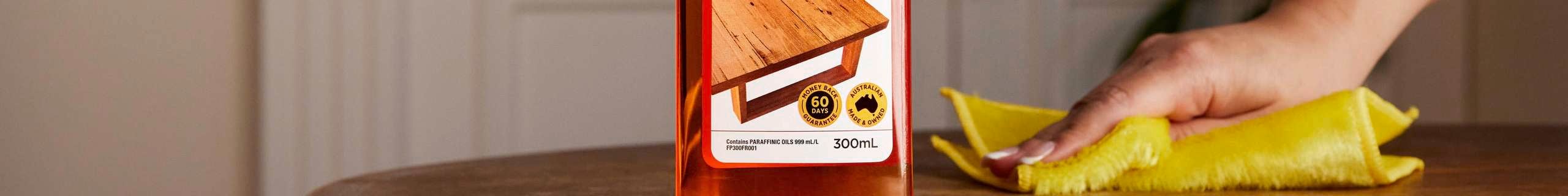 A person's hand holding a cloth against a wooden table with a bottle of O'Cedar furniture polish on it.
