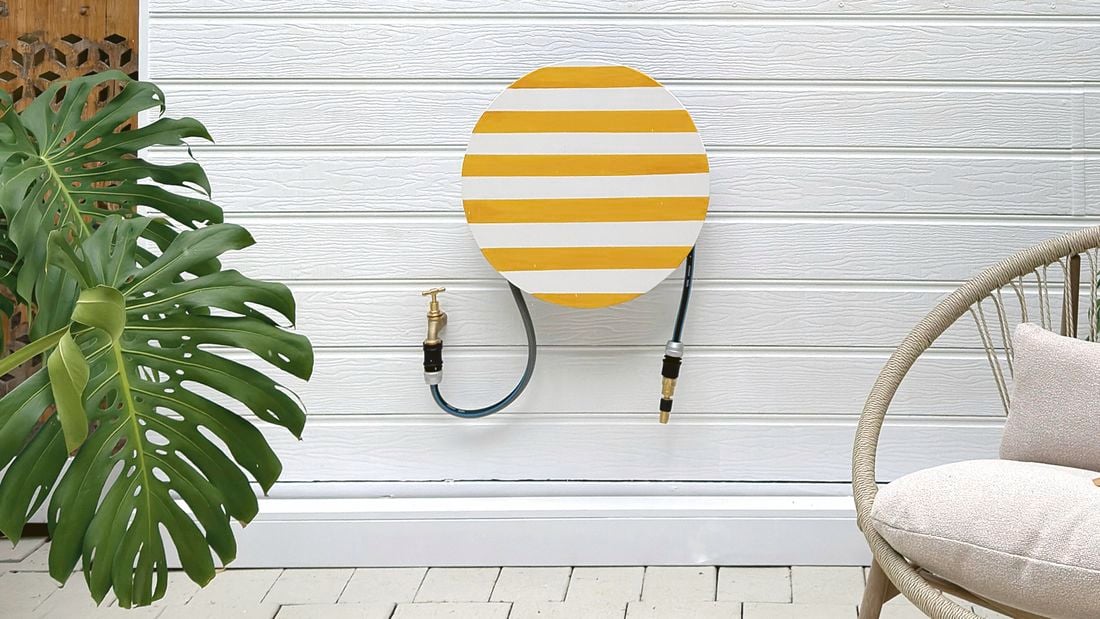 A garden patio with a large potted Monstera plant, a striped hose hanger cover on a white wall, and a woven outdoor chair