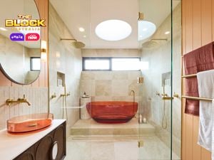 Modern bathroom with amber tub in glass shower, beige tiles, dual overhead showerheads, amber basin with round mirror, and gold towel rack. 