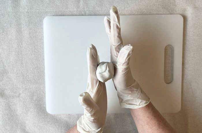  A pair of hands wearing white gloves shapes white clay into a ball over a white cutting board.