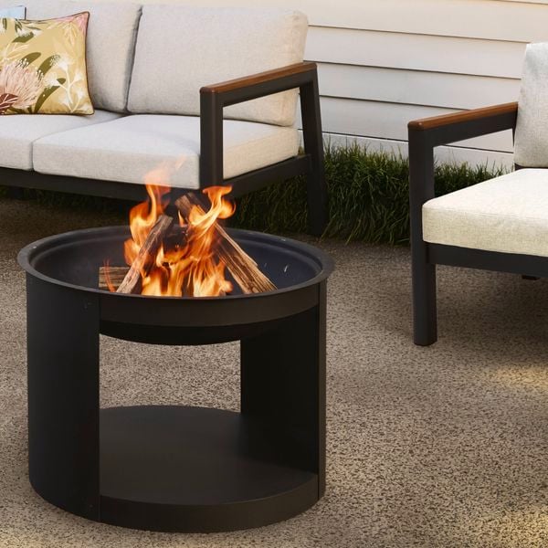 Black round outdoor fire pit with a visible flame, placed on a concrete patio and surrounded by modern outdoor lounge seating with light cushions and decorative pillows.