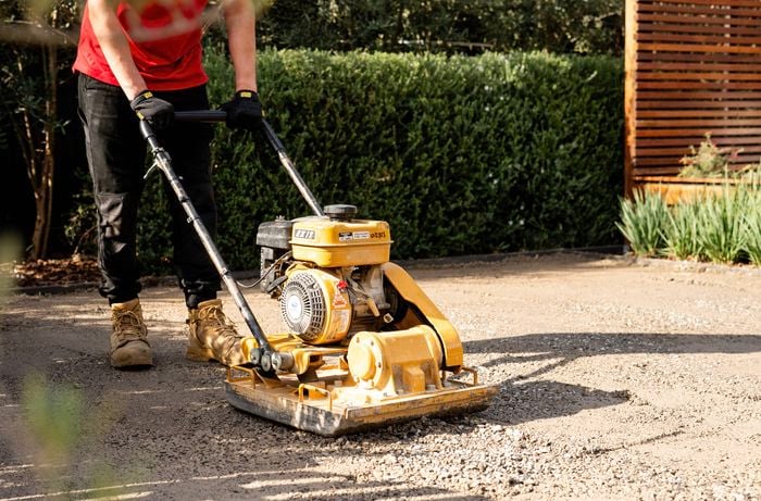 A person wearing gloves and work boots is using a compactor to compress the road base on the ground