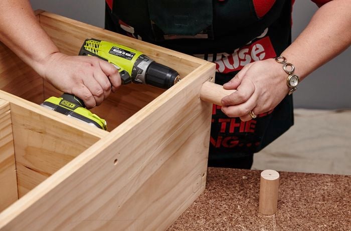 Wooden dowel legs being added to the underside of a wooden storage bench with a power drill