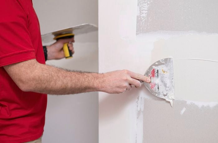 A person in a red T- shirt is applying plaster to a wall while holding a plastering trowel in one hand and a putty knife in the other