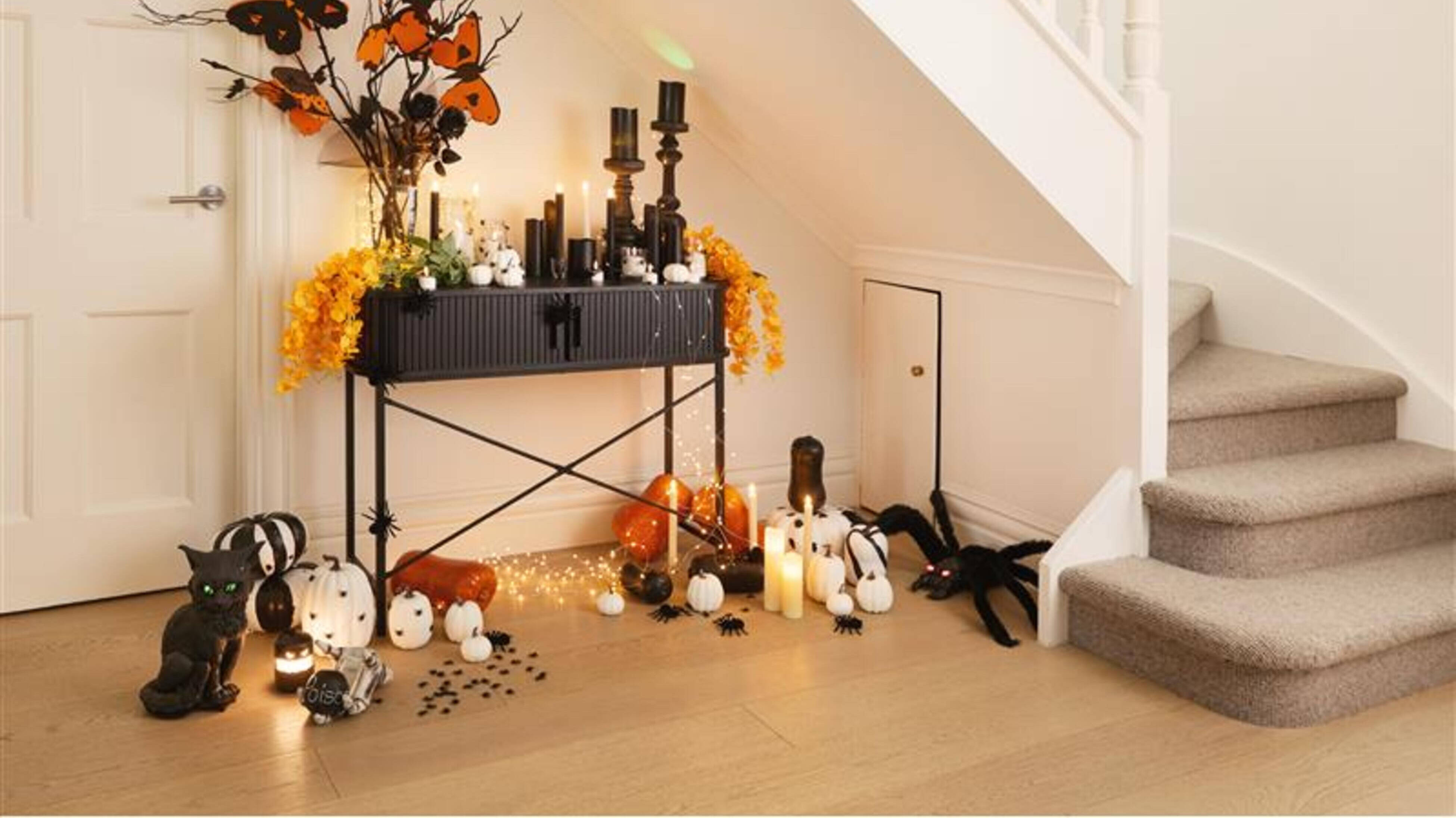 A Halloween-themed display under a staircase featuring black and orange decorations, candles, pumpkins, spiders, and a black cat figurine.