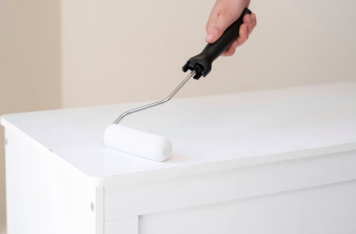 A person is applying primer to a timber toy box using a paint roller to create a solid base for painting.