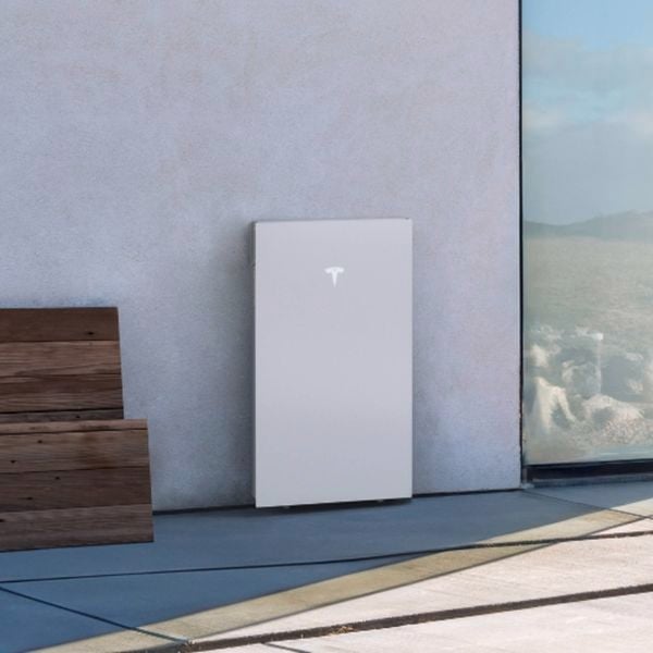 A Tesla Powerwall 3, rectangular in shape and white in colour, cleanly installed on a garage wall of a home, mounted above the ground and with cabinetry alongside.