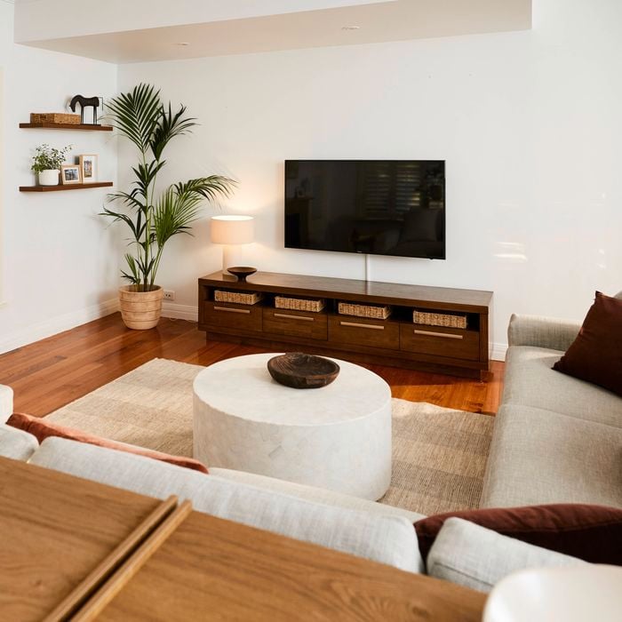 Light-filled living room with wooden floors, a wall-mounted TV above a low timber cabinet with baskets, a round coffee table on a rug, neutral sofas, indoor plants, and minimalist wall shelves.