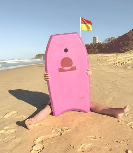 Child sitting on the sand hidden behind a pink bodyboard with a surf lifesaving flag behind.