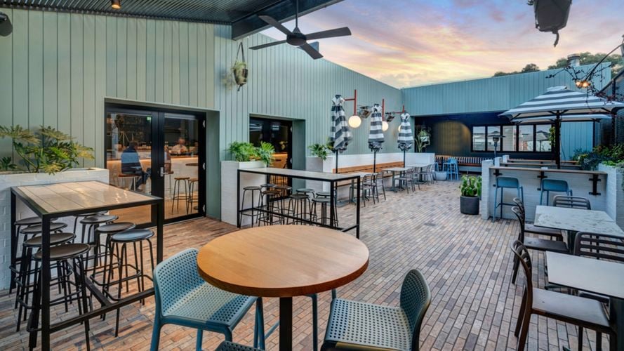 Outdoor patio of a restaurant or café with mixed seating including bar stools and regular chairs, potted and hanging plants, ceiling fans, large umbrellas, and a colorful sunset in the background.