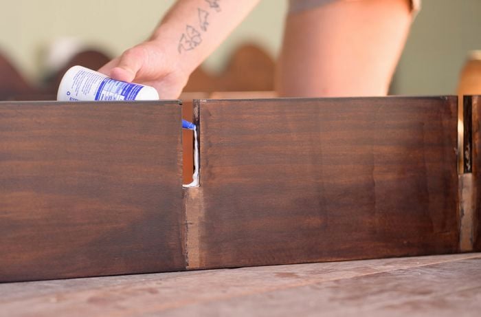 A person is applying wood adhesive into a notch on a stained wooden panel, preparing it for assembly for the bookshelf.