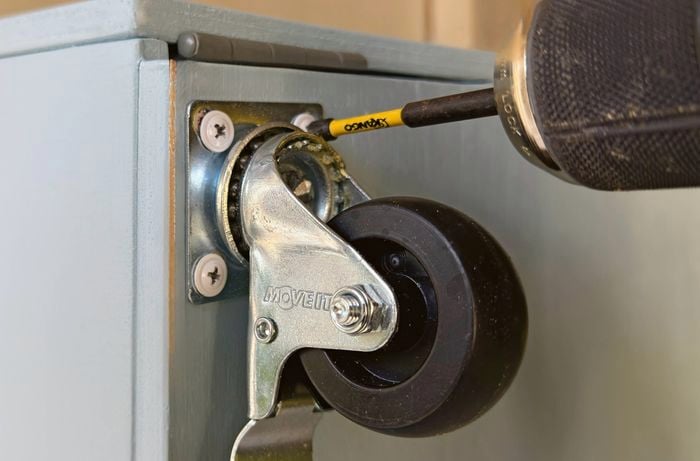 A drill attaching a castor wheel to a cabinet.