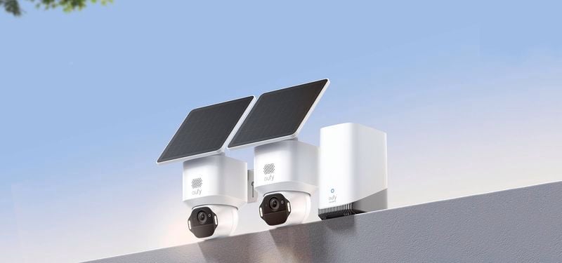 Two white eufy solar security cameras with tilted black solar panels mounted on an outdoor wall, with a storage or base unit beside them under a clear blue sky.
