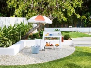 A white drink trolley sits in a backyard, set up with fruit, glasses and drinks, next to a red and white shade umbrella.