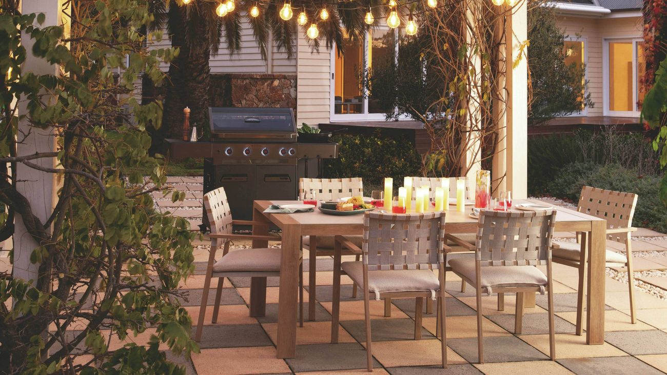 Outdoor dining area beneath a pergola draped with foliage and illuminated by string lights, with a table, woven chairs, and a barbecue in the background.