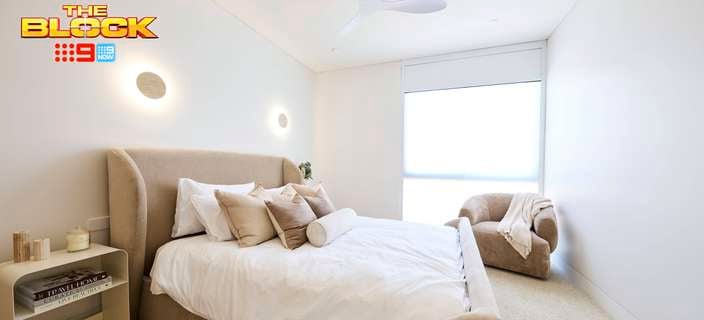 A furnished bedroom with a bed in it, a side table, a chair and carpet on the floor with The Block logo on the image.