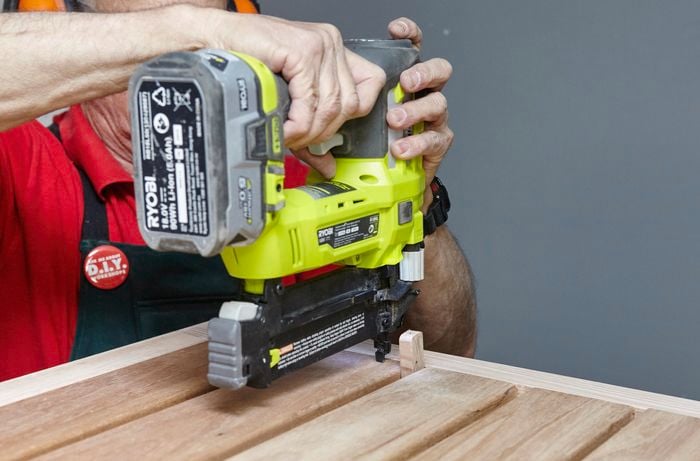 The slats of a wooden chair's seat being nailed into place with a nail gun, with a scrap piece of timber used as a spacer
