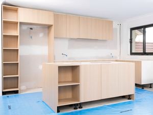 The image shows a kitchen under renovation, with new light wood cabinetry being installed and blue protective sheets covering the floor