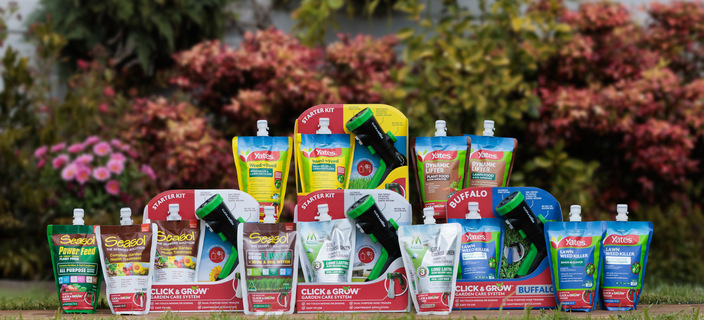 Garden care products and hose-end applicator kits displayed on a lawn with flowering garden beds in the background.