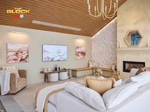 A modern living room with a high wooden ceiling and a large chandelier. The space includes a white and beige sectional sofa with pillows, an armchair draped with a blanket, and two small round stools. A fireplace on the right is framed by stacked firewood, candles, and an octagonal mirror above. The back wall features two paintings flanking a central TV displaying flowers. The left wall is clad in textured stone.