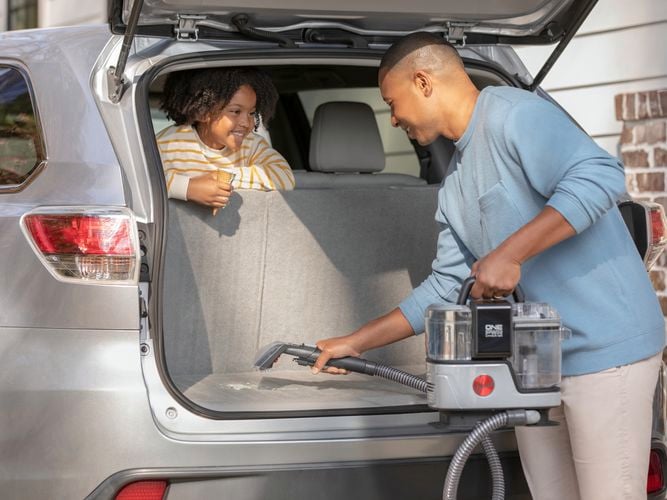 A man and child seated in the rear of a car, engrossed in conversation, cleaning the boot upholstery with a Hoover Clean Slate Pot Washer.