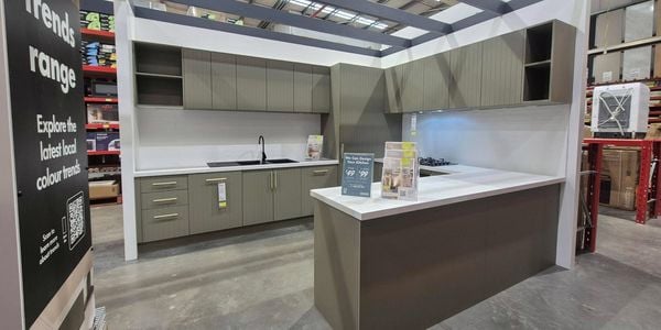 Kitchen showroom display inside a Bunnings Warehouse store, featuring grey cabinetry, white benchtops, a black sink, overhead storage cupboards and a central island beneath a decorative pergola-style frame. Informational displays and brochures are positioned on the benchtop.
