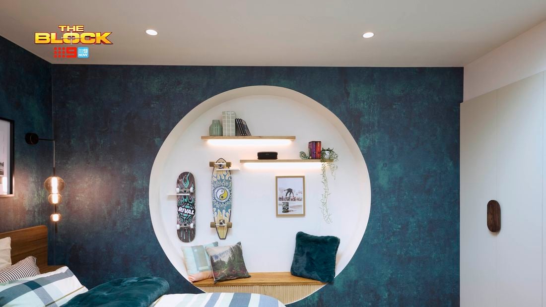 Blue bedroom with a double bed, textured blue wallpaper and blue checked linen. The bed is on a brown checked rug and there is a circular recess in the wall with a bench seat and clothing hooks.