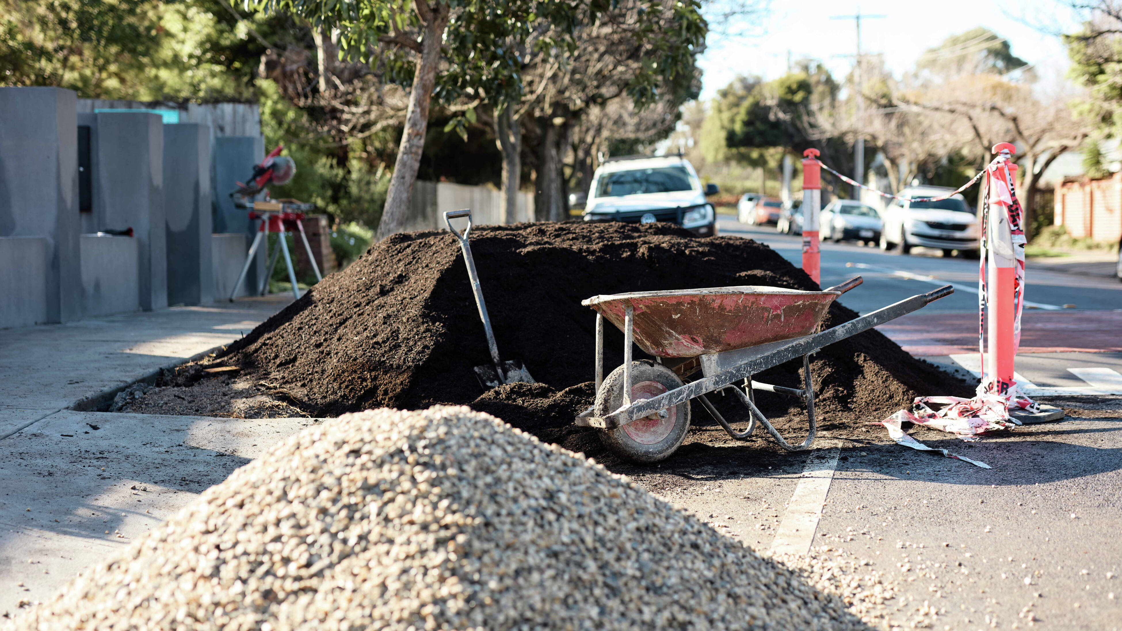 A wheelbarrow, a shovel, and large piles of soil and gravel on a suburban street during landscaping work