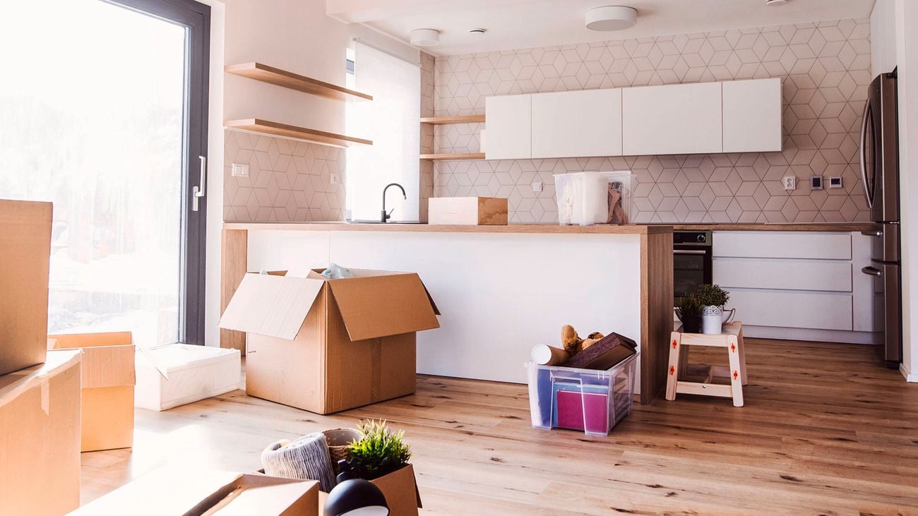 Modern kitchen with open cardboard moving boxes on the floor, plastic tubs, and a small wooden stool