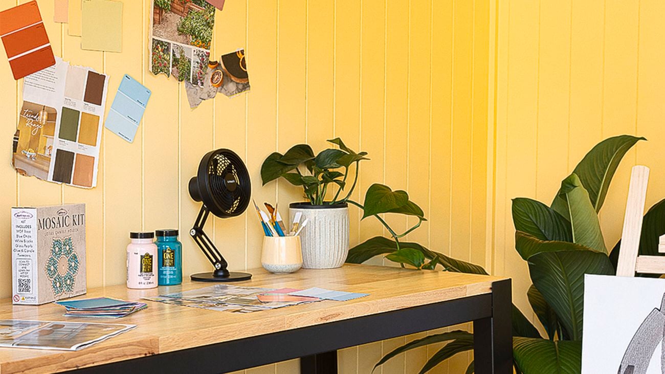 Close up of the interior of a shed showing yellow-painted wall panelling and a timber desk with metal legs, with a pot plant, paint swatches, a fan, craft paints and a mosaic kit on top. A large-leafed plant and an easel are off to the side.