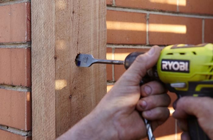 A person drilling hole in timber post against a brick wall