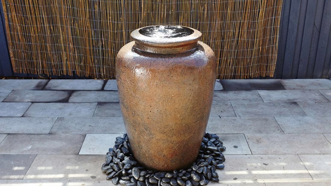 A large brown stone pot with bubbling water in a paved outdoor area with pebbles